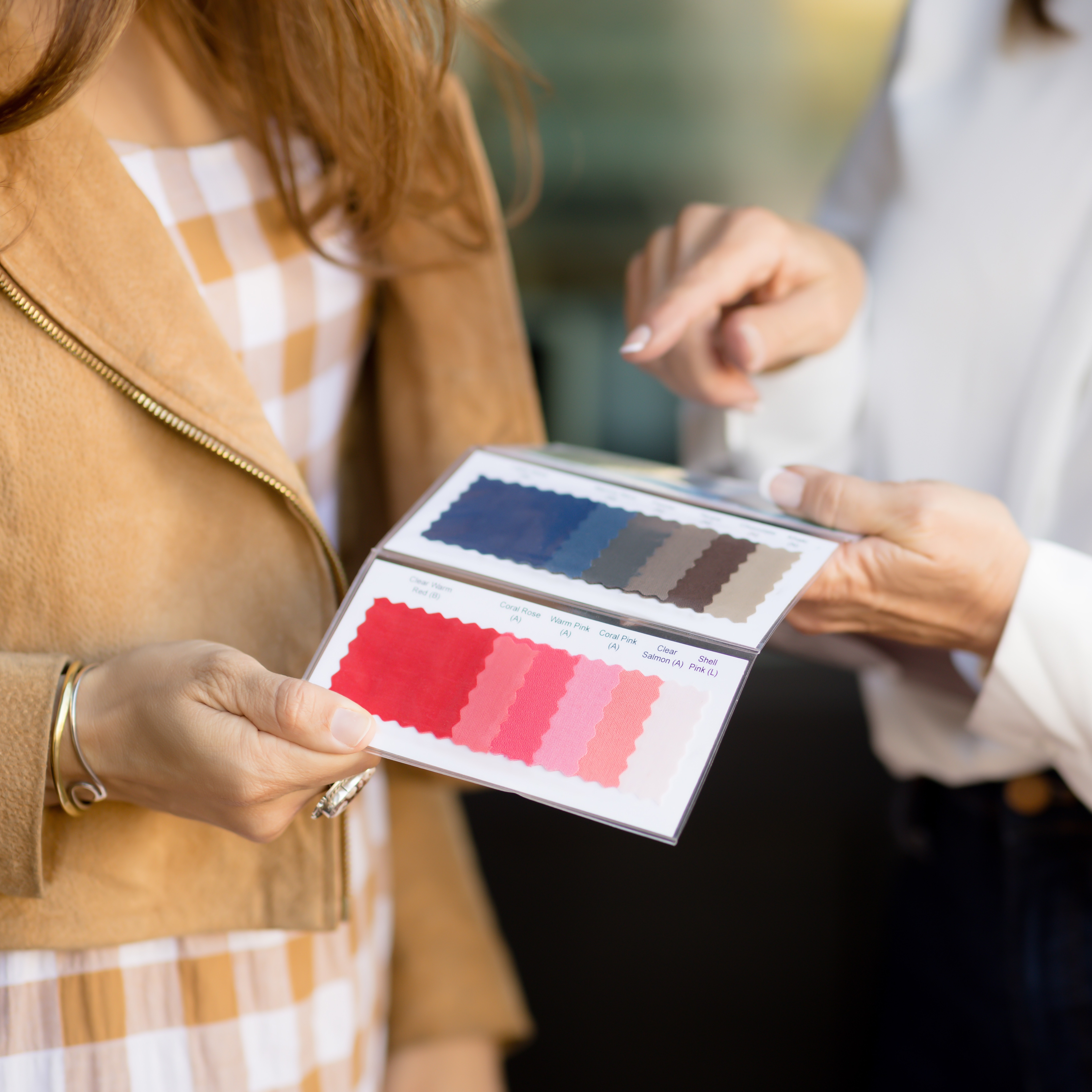 Two people examining a color swatch book. The person on the left, wearing a brown jacket, holds the book open to reveal a perfect palette of fabric samples. The person on the right points at the book with one finger, as if engaging in personal color analysis.