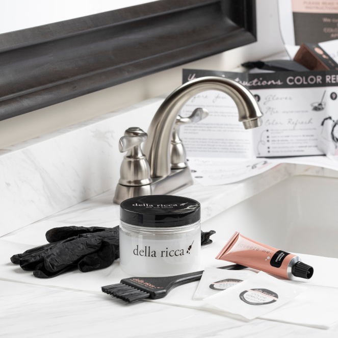 On the bathroom countertop sits a custom at-home hair color system, featuring a hair dye kit with premium ingredients: a jar labeled "della ricca," a tube of color, gloves, brush, instructions, and two conditioner packets. In the background are a faucet and mirror.