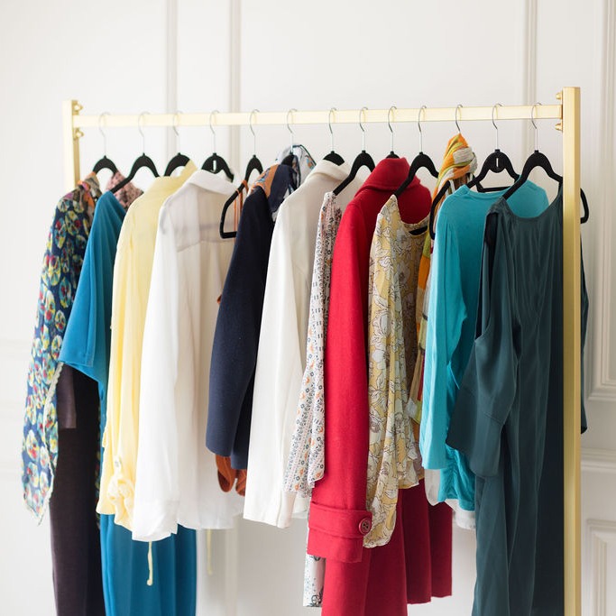A gold clothing rack showcases a vibrant collection of garments, including dresses, blouses, and jackets in hues like red, blue, yellow, and floral patterns. Perfectly complementing the white wall backdrop, each piece awaits your personal stylist to help refine your style with ease.