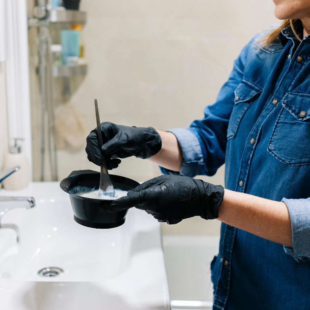 A person wearing black gloves and a denim shirt holds a black bowl of at home hair color with a stirring tool over a sink in the bathroom, perfecting their at-home hair color. Various toiletries are visible in the background, hinting at personalized formulas crafted with care.