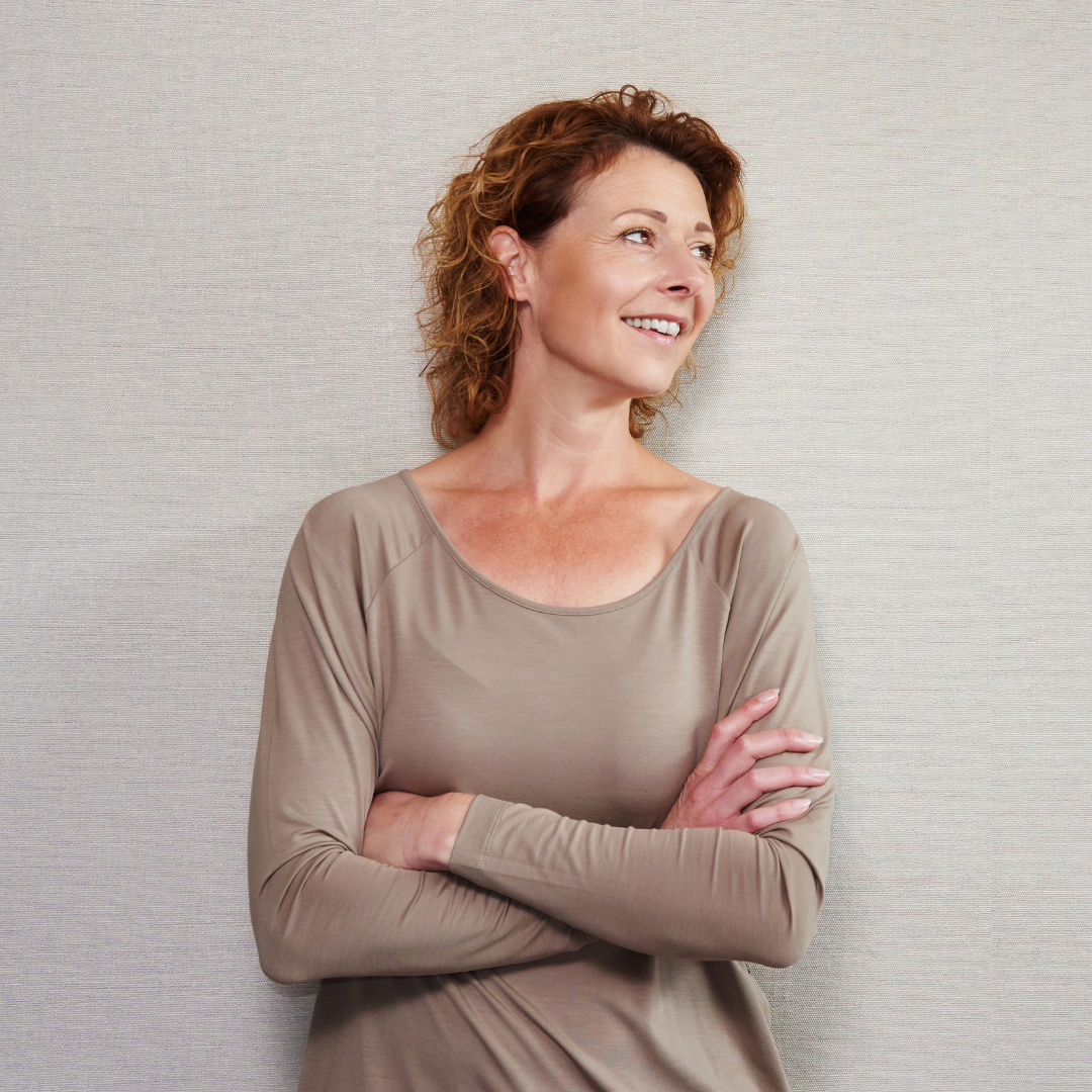 A woman with curly hair stands against a light-colored wall, smiling and looking to the side, embodying the confidence of custom at-home hair color. She is wearing a beige long-sleeve top and has her arms crossed.